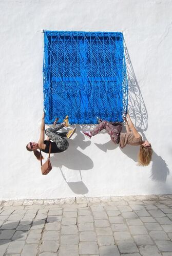 Two people who appear to hang from a blue window grille on a white wall